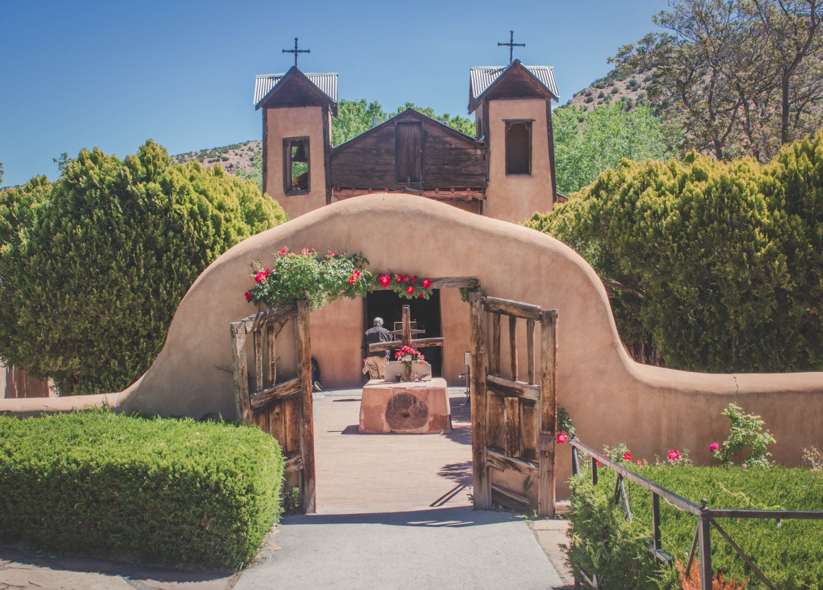 Gorgeous entrance to El Santuario De Chimayo, with flowers and greenery all around. The brick is a pretty shade of brown. And the gates look rustic. 